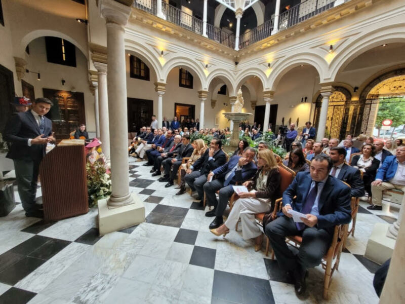 Conferencia en un palacio histórico con columnas y arcos, asistentes en trajes formales, y un escenario con microfonos.