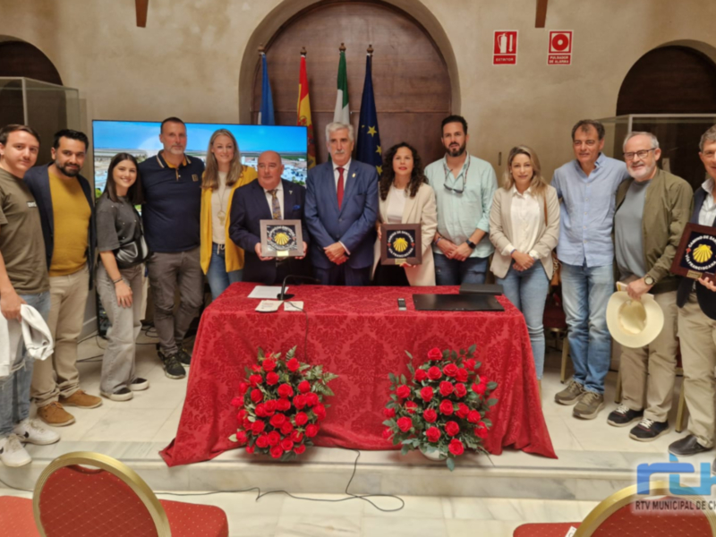 Una reunión formal en un salón con una mesa cubierta de tela roja y flores rojas. Los asistentes sostienen placas con insignias y sonríen para la cámara. En el fondo, se pueden ver banderas y un televisor con una imagen de un puente.