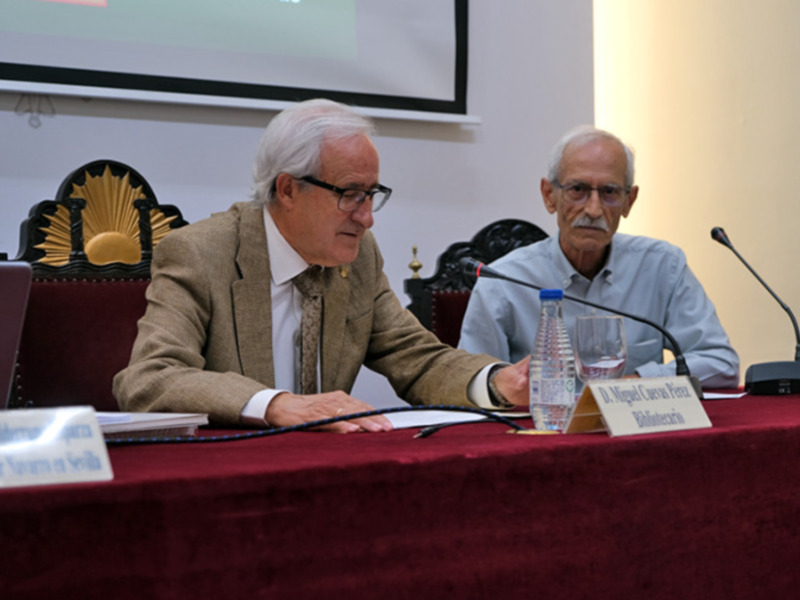 Dos hombres sentados en una mesa con micrófonos, participando en una reunión o conferencia.