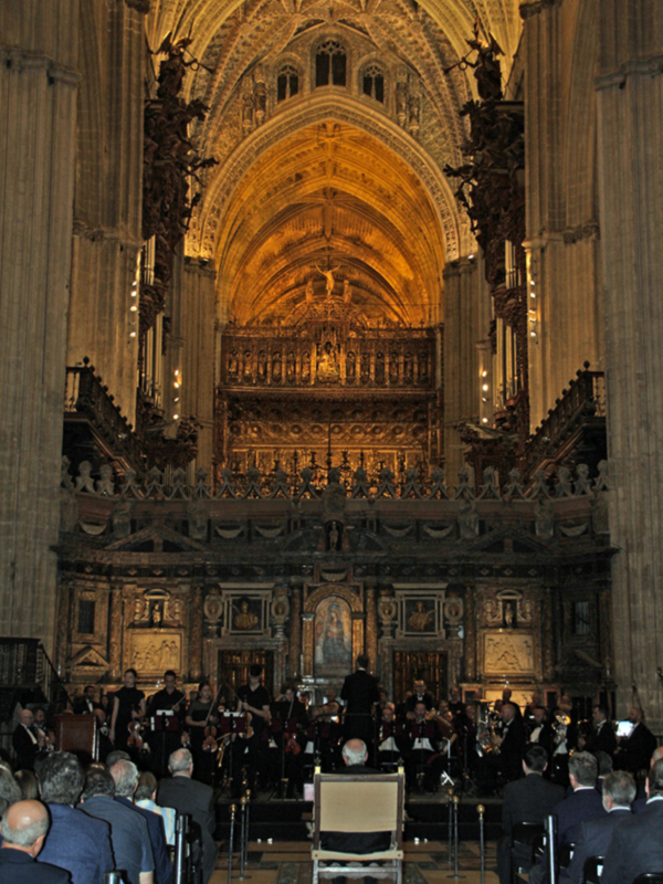 Orquesta y coro en un concierto en una catedral gótica, con arcos y detalles artísticos en el fondo.