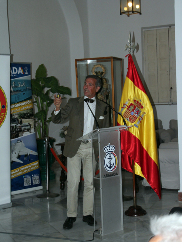 Hombre hablando en un evento con bandera española y carteles de la Armada en el fondo.