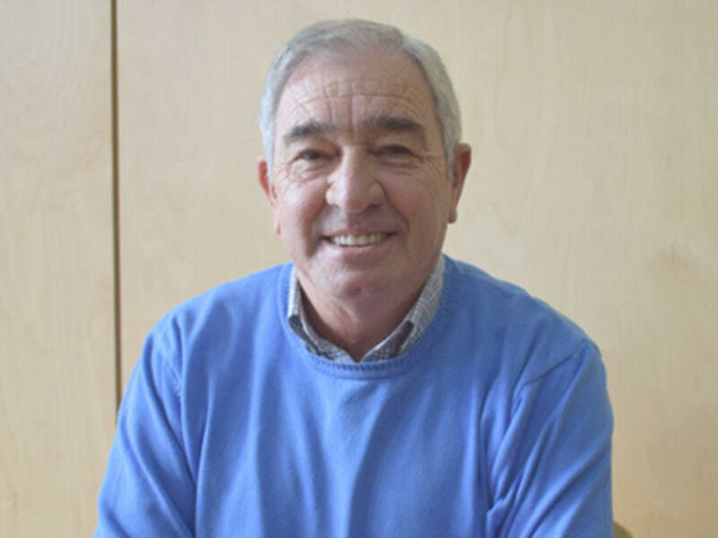 Retrato de hombre sonriente, vestido con suéter azul, fondo de madera clara.