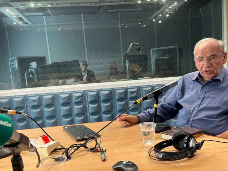 Un hombre en un estudio de radio, sentado frente a un micrófono y un teclado, con auriculares y una taza de agua en la mesa. El estudio tiene un fondo de cristal y paneles acústicos azules. El hombre parece estar en plena transmisión.