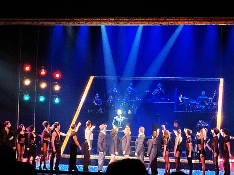 Escenario de un espectáculo con luces azules y rojas, bailarines en formación y orquesta en el fondo.