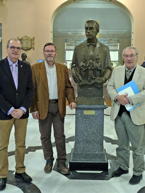 Tres hombres posan frente a un busto en un ambiente formal, probablemente en un evento de homenaje o reconocimiento.
