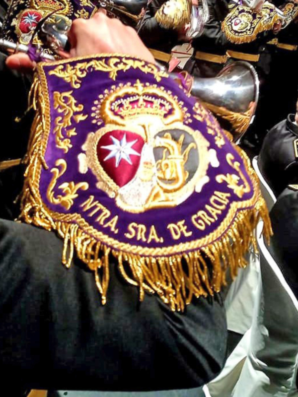 Detalle de un escudo con el monograma de la Virgen de Gracia, adornado con franges dorados y un fondo de color púrpura.