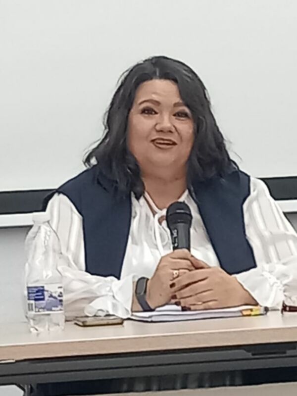 Mujer hablando en un evento público, sosteniendo un micrófono y sentada en una mesa con un botella de agua y un teléfono móvil.
