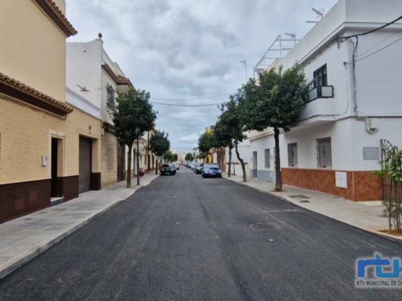 Calle tranquila con casas de fachadas blancas y amarillas, árboles y coches estacionados en Chipiona, España.