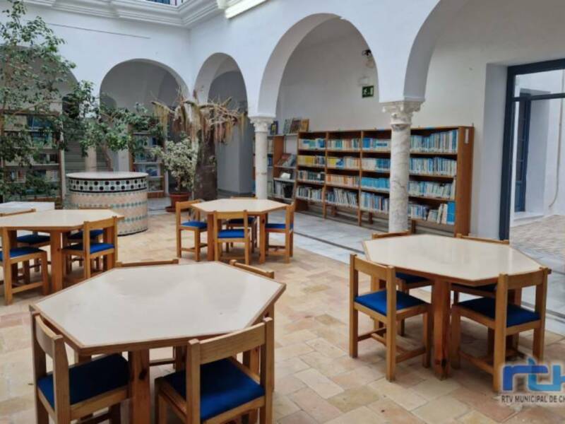 Interior de una biblioteca con arcos blancos, mesas y sillas, y estanterías de libros.