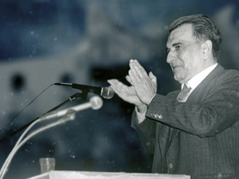 Persona en traje hablando en un podio con micrófono, fondo borroso