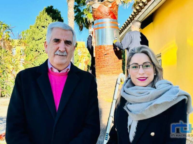 Dos personas posan frente a una pared amarilla con un hombre trabajando en el fondo.