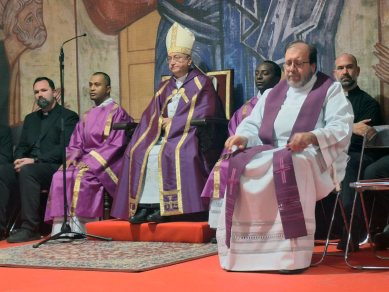 Celebración religiosa con sacerdotes en trajes litúrgicos y un fondo con pinturas religiosas.