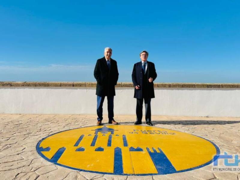 Dos hombres en trajes frente al logo del Puerto de Sevilla, con el cielo azul como fondo.