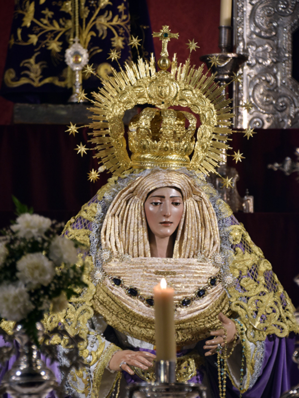 Imponente imagen de la Virgen María con manto dorado y corona de estrellas, en un entorno religioso con velas y flores.