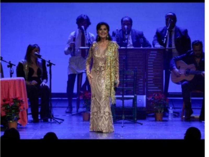 Una cantante en escenario con un vestido dorado y flores, acompañada por músicos en el fondo.