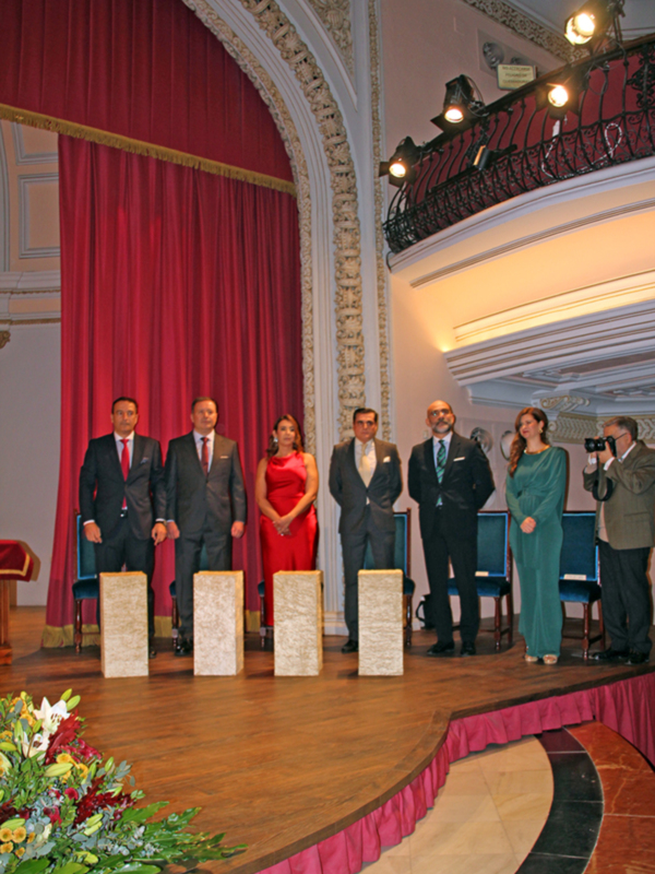 Oficiales y personalidades en un evento formal en un salón decorado con cortinas rojas y luces de techo.