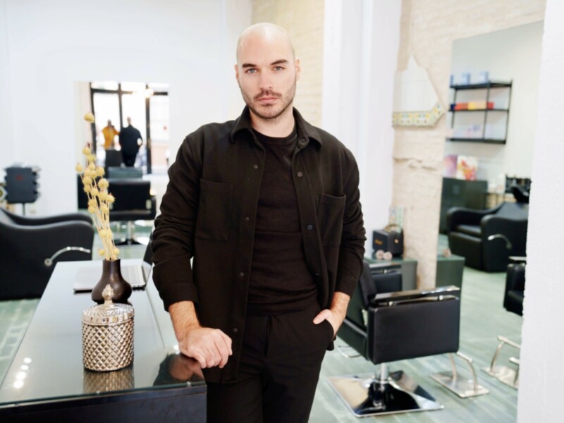 Man standing in a stylish salon, dressed in black, with a modern interior.