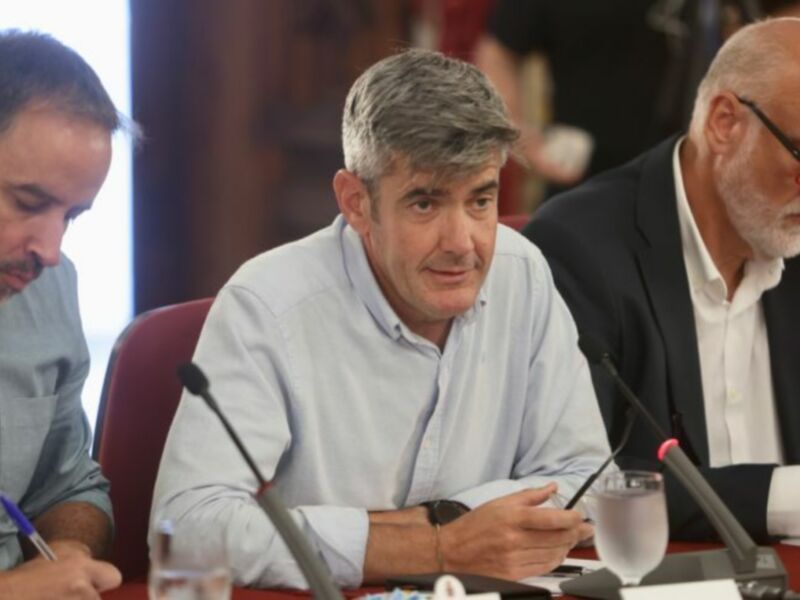 Reunión de trabajo en una mesa con micrófonos y documentos, tres hombres sentados.
