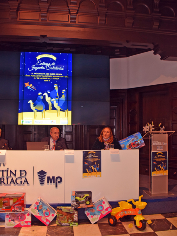 Presentación de juguetes solidarios en un evento público, con un fondo de pantalla que muestra animales de la noche y un mensaje informativo sobre la donación de juguetes.