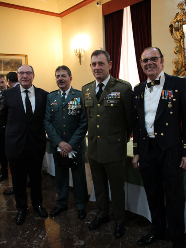 Oficiales militares y civiles en una ceremonia formal en un salón decorado con cortinas rojas y lámparas de pared.