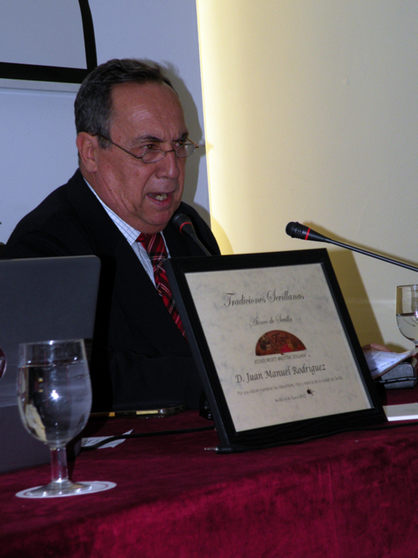 Hombre hablando en un evento con un diploma de 'Tradiciones Sevillanas' en primer plano.