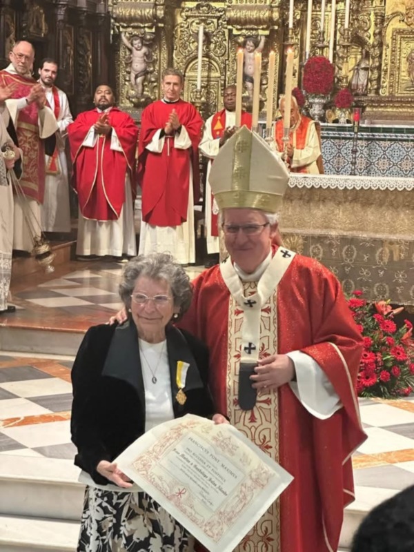 Celebración religiosa con un sacerdote y una mujer recibiendo un certificado en un ambiente eclesiástico.