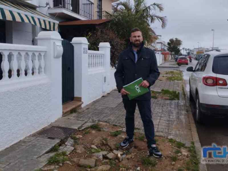 Un hombre con una chaqueta negra y una chaqueta verde en la mano, posando frente a una casa blanca con un toldo amarillo y verde. El hombre tiene barba y está en una calle con un coche blanco estacionado a su lado.