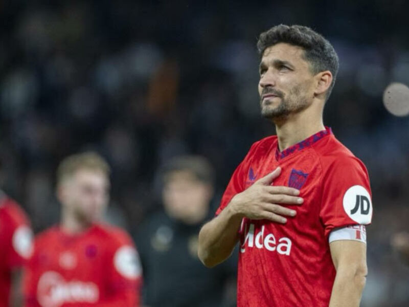 Jugador de fútbol con camiseta roja y brazalete en la manga, posando con la mano en el pecho.