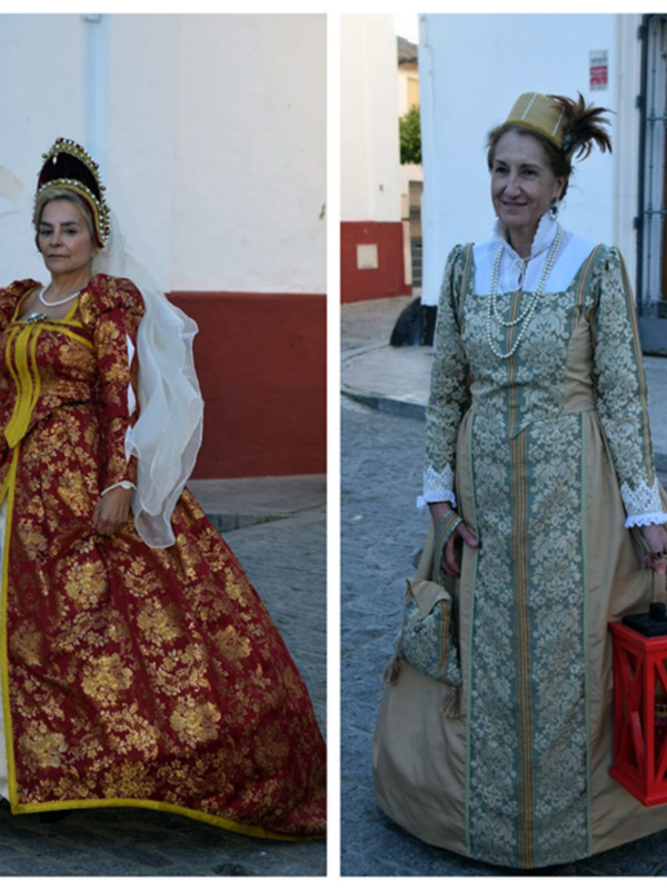 Mujeres vestidas con trajes tradicionales españoles, uno en rojo y dorado y otro en verde y beige, en una calle de pueblo.