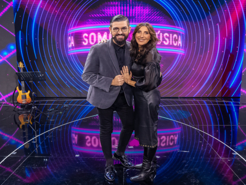 Presentadores en escenario de 'Somos Música', con fondo futurista y guitarra en el escenario.