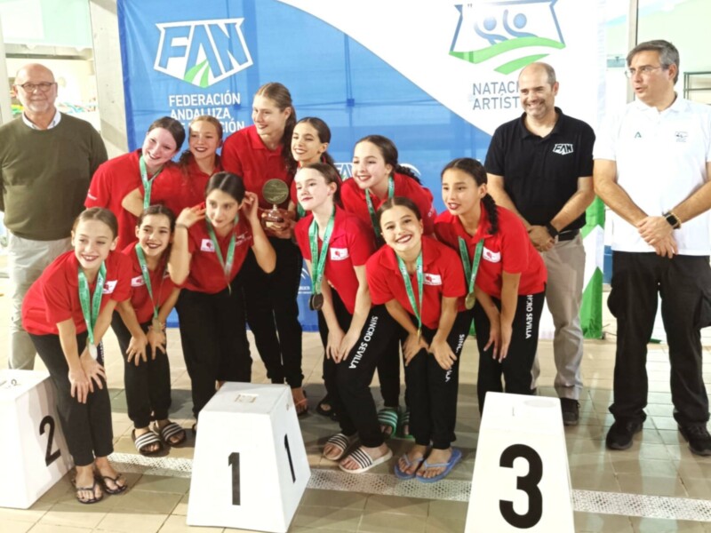 Equipo de natación artística posando con medallas y trofeo en el podio de una competición de la Federación Andaluza de Natación, con el logo de la FAN y el de Natación Artística en el fondo.