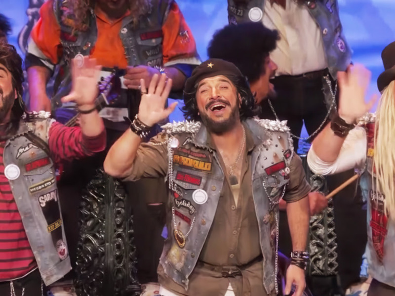 Un grupo de personas vestidas de manera casual y con accesorios como chaquetas con parches y collares, celebrando con las manos en el aire en un escenario con luces azules en el fondo.