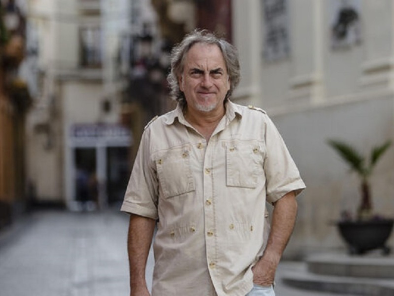 Hombre con camisa beige sonriendo en una calle de edificios antiguos.