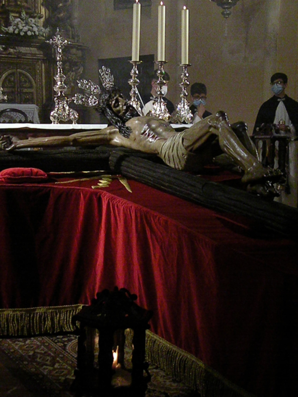 Estatua de Cristo en la cruz en un altar con velas encendidas y una mantel rojo. A la derecha, dos personas con vestimentas de oficiales de la policía, mirando hacia la estatua. El ambiente es religioso y ceremonial.