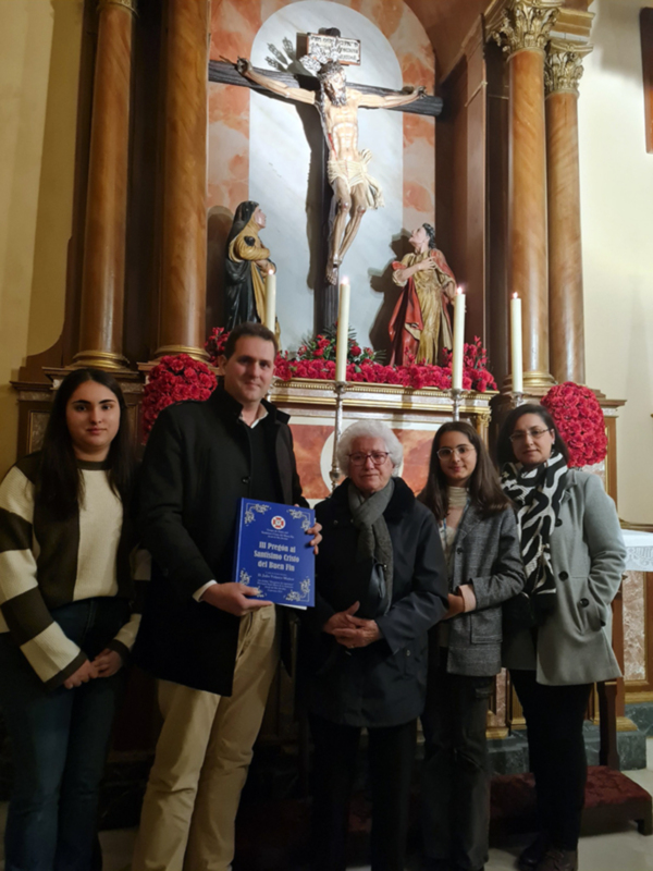 Grupo de personas posando con un libro en un altar con estatuas religiosas y flores rojas.