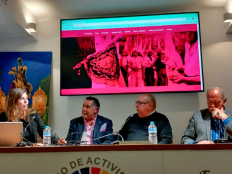 Una conferencia de prensa en un salón con tres personas sentadas en una mesa, con un televisor en el fondo mostrando imágenes de una procesión religiosa.