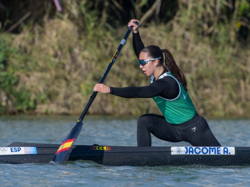 Una atleta de canotaje en acción, con su canoa marcada con el logo de los Juegos Olímpicos de París 2024. La atleta usa una camiseta verde y negro con el logotipo de su equipo, y una canoa con la bandera de España y el nombre de su club, Jacomea.