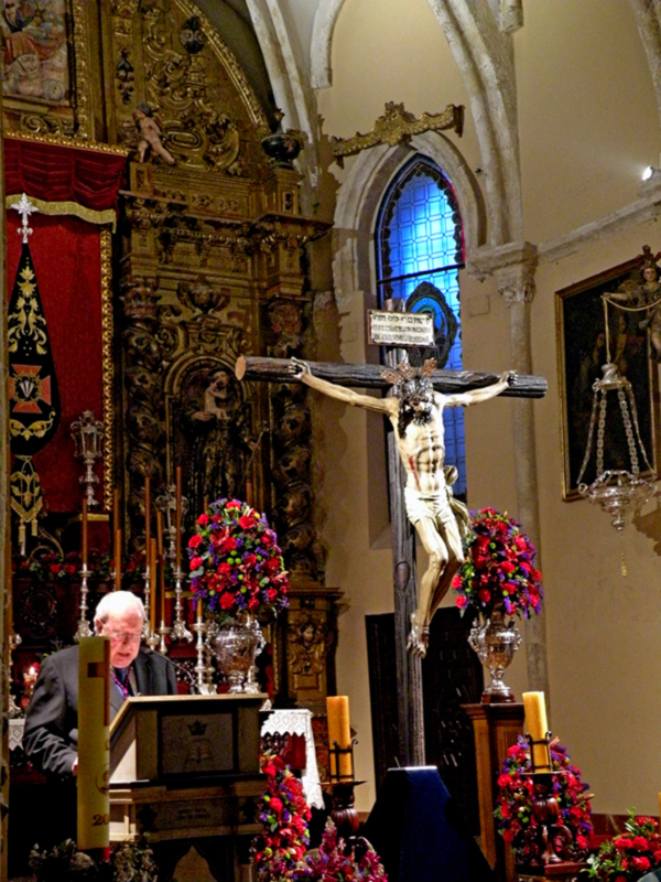 Interior de una iglesia con un altar ornamental, un crucifijo y flores en el centro, y un hombre hablando desde un púlpito.
