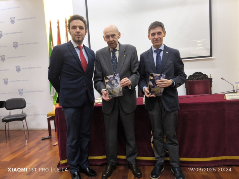 Presentación de un libro en el Ateneo de Sevilla, con tres personas posando junto al libro. El fondo muestra el escudo del Ateneo y la silla presidencial. La fecha y hora de la imagen se encuentran en la parte inferior derecha.