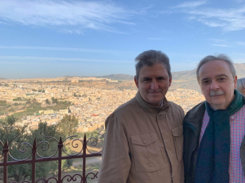 Dos personas frente a una vista panorámica de una ciudad con edificios y montañas en el horizonte, bajo un cielo azul con algunas nubes. La imagen sugiere un momento de descanso o contemplación en una ubicación con vistas panorámicas.