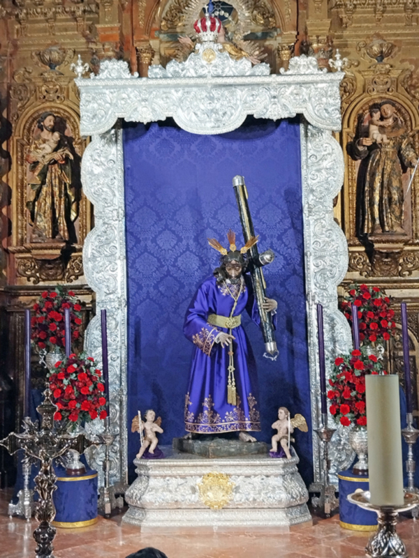 Elegante estatua religiosa en un altar con adornos dorados y flores rojas, rodeada de detalles artísticos y una profunda textura azul en el fondo.