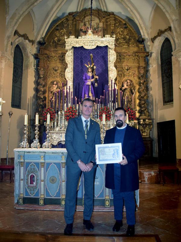 Celebración religiosa en un templo con dos personas posando frente al altar, uno sosteniendo un certificado.