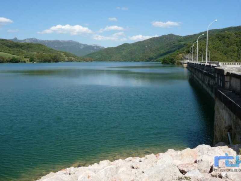 Lago con presa y montañas al fondo bajo un cielo azul claro.