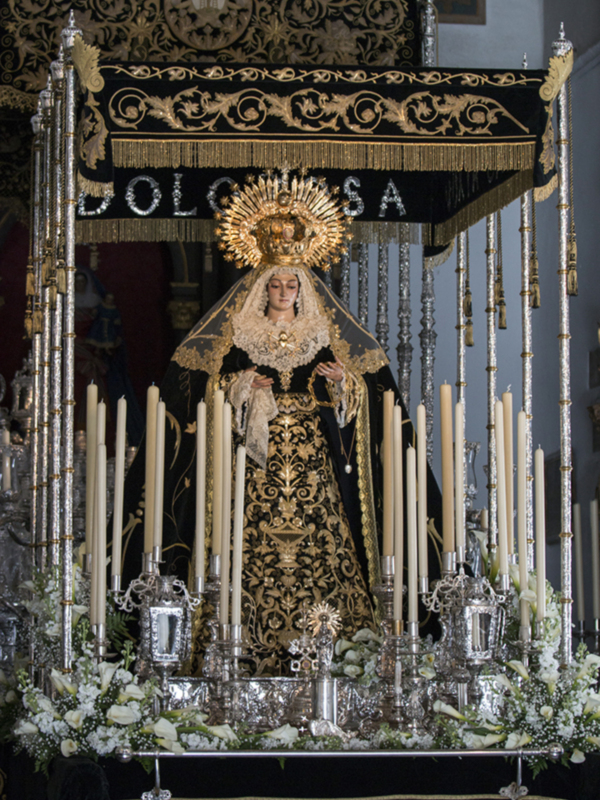 Imaginación: Una imagen de una Virgen María vestida en trajes tradicionales, rodeada de velas y flores, posiblemente en un altar de una iglesia católica.