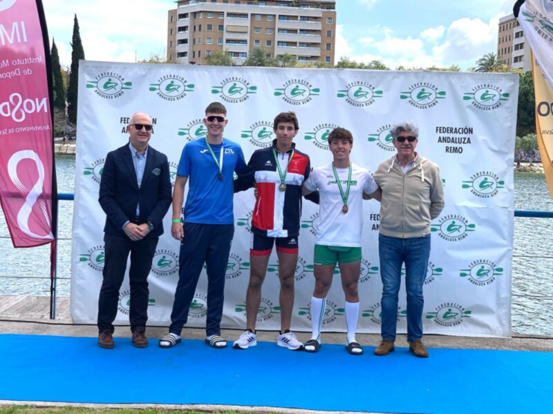 Tres atletas posan en un podio junto a un oficial de la Federación Andaluza de Remo, celebrando una victoria en un evento deportivo.