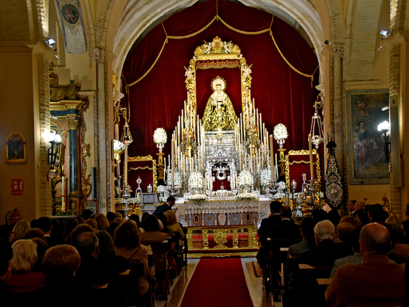 Interior de una iglesia con una imagen religiosa en el altar, rodeada de velas y adornos, mientras una multitud de personas asiste a una ceremonia religiosa.