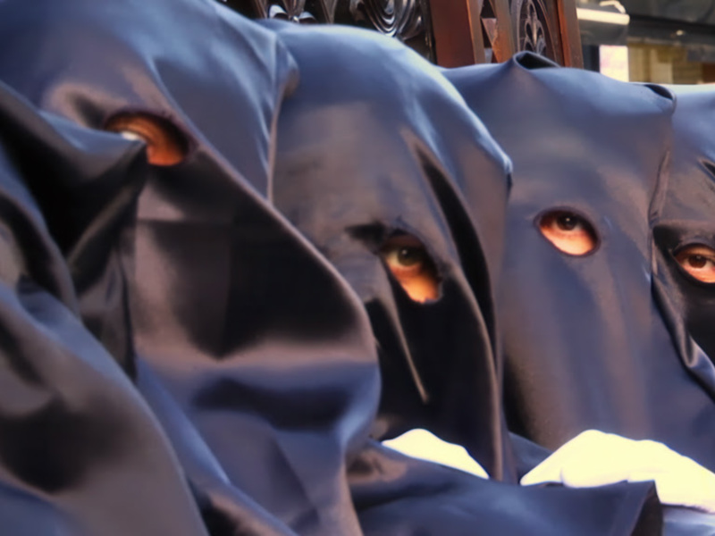 Procesión con penitentes en capas negras y capuchas con ojos visibles.