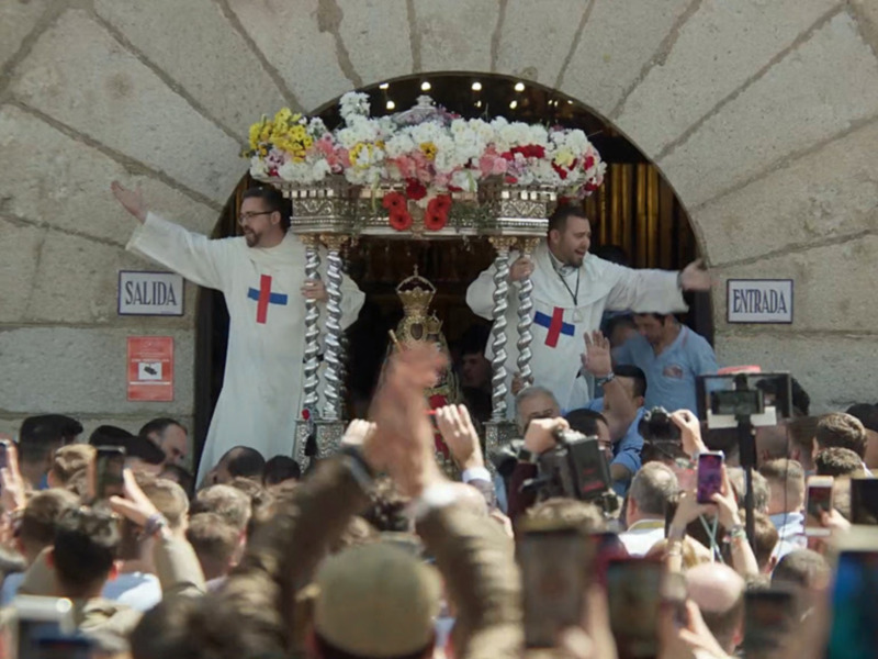 Procesión religiosa con sacerdotes portando un estandarte ornado con flores y cruz, rodeados por una multitud entusiasta.