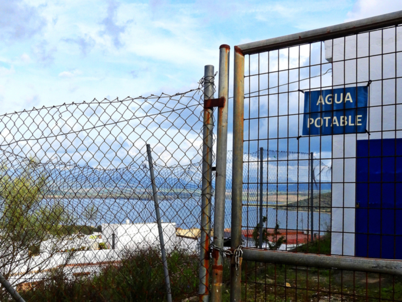 Fence with 'AGUA POTABLE' sign, overlooking a body of water and industrial buildings.
