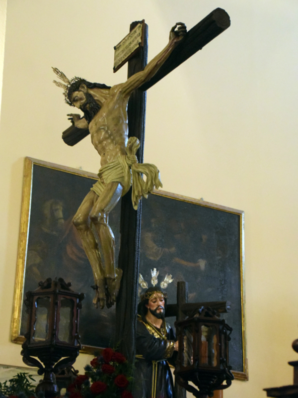Escultura de Cristo en la cruz en un entorno religioso, con detalles artísticos y flores rojas en el suelo.